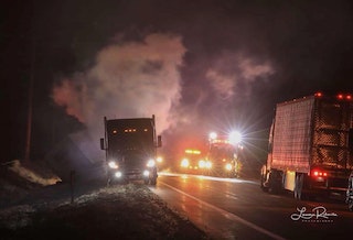Night scene with trucks on road, smoke in the air, and emergency vehicle lights.
