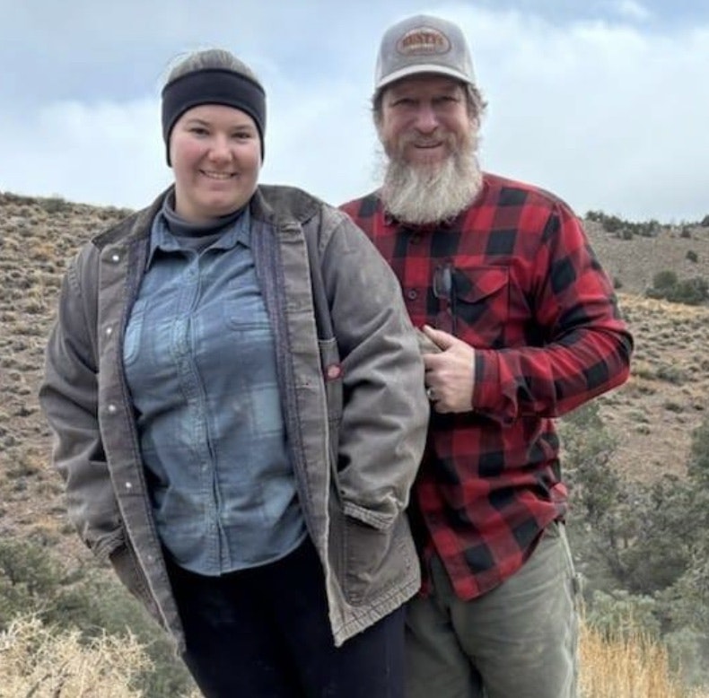Two people outdoors, dressed warmly in jackets and beanie, standing on a grassy hill.
