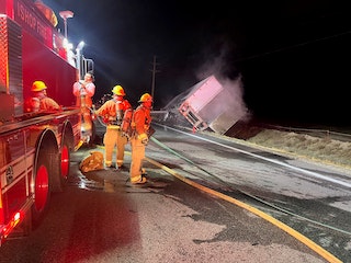 Firefighters beside a fire truck at night, with an overturned vehicle in the background.