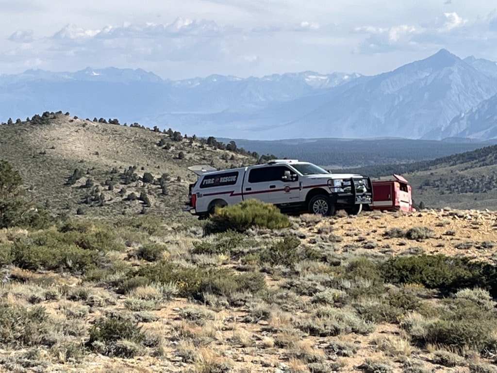 A fire rescue vehicle is parked in a rugged, shrubby terrain with mountains in the background.