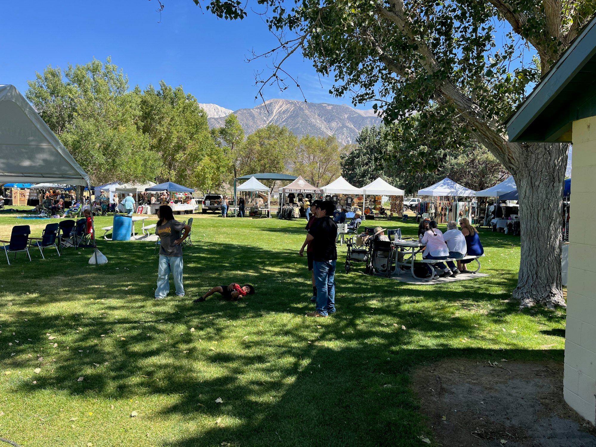 Outdoor event with people, tents, and picnic tables on a grassy area with trees and mountains in the background.