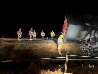 A group of people near an overturned truck at night, illuminated by headlights, possibly emergency responders at an accident scene.