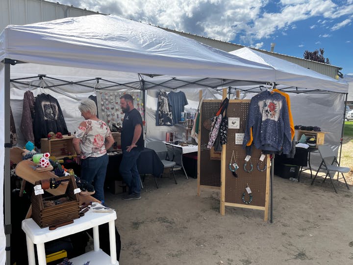 Outdoor market stall with clothes, accessories, and decorations on display. Two people browsing under a white canopy.