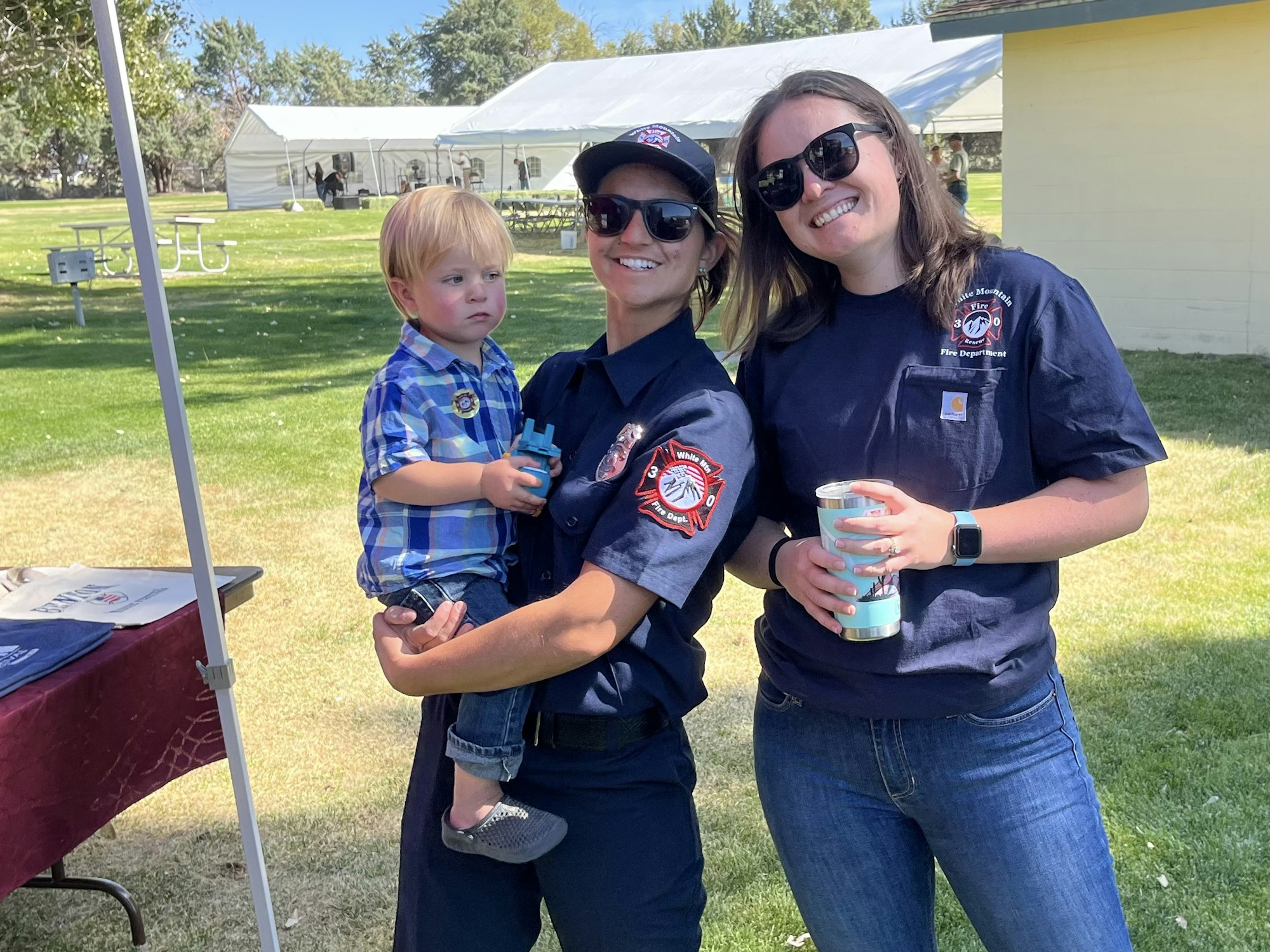 Two women in firefighter shirts and a toddler at an outdoor event.