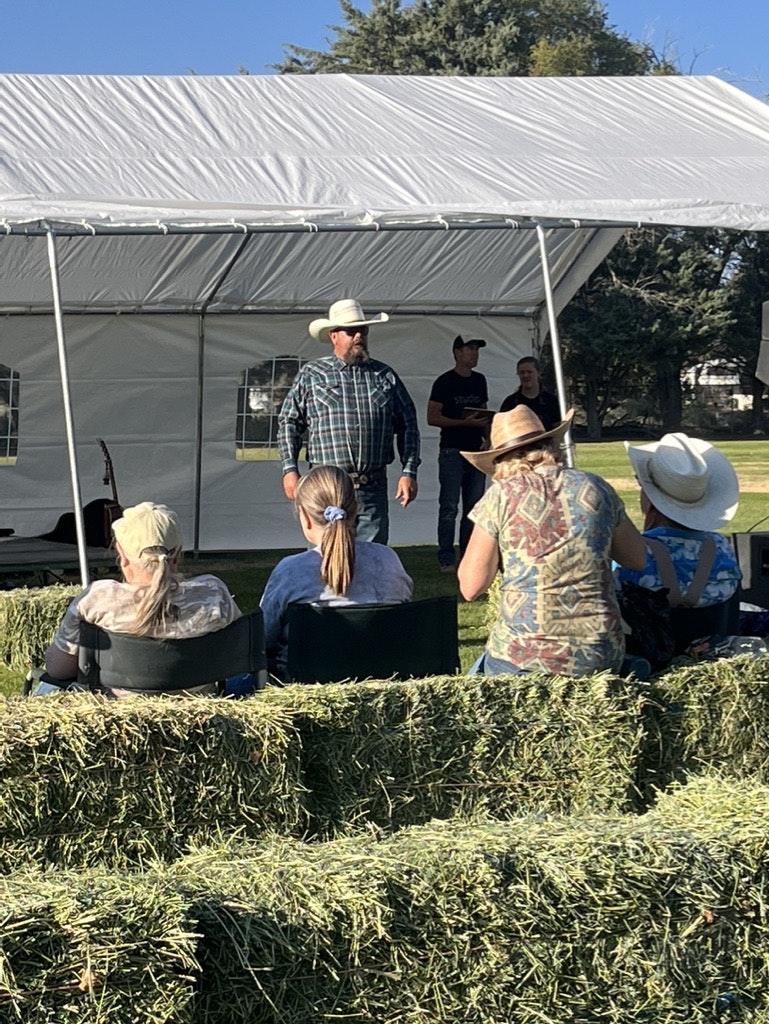 People at an outdoor event, sitting on hay bales, with a man standing under a tent.