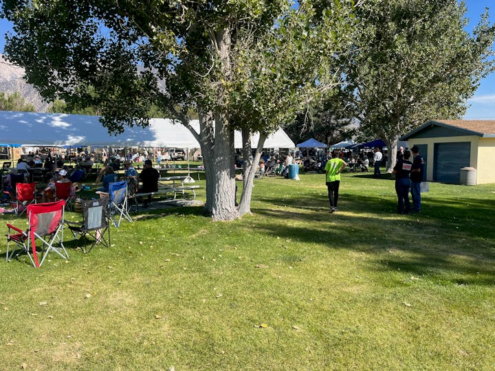 Outdoor gathering with people sitting under tents and around tables in a park, surrounded by trees and open grass.