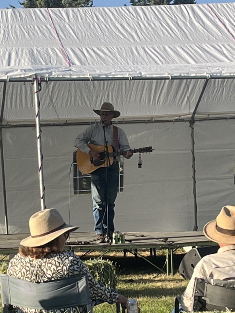A person playing guitar on stage at an outdoor event with an audience in hats listening.
