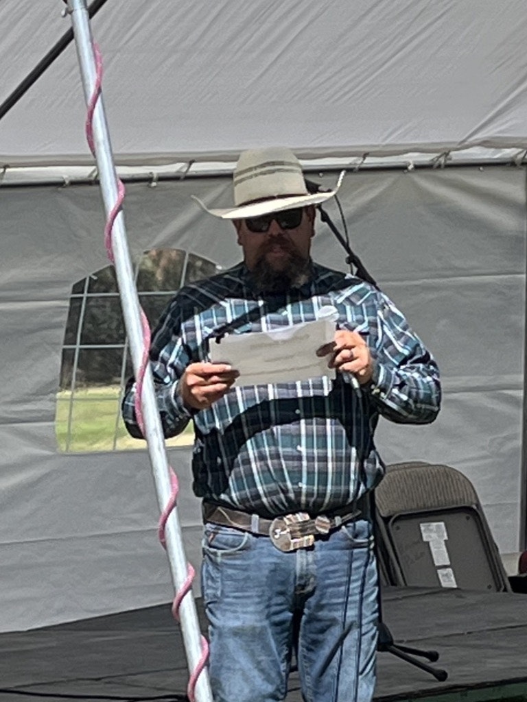 Man with a cowboy hat reading a paper on a stage inside a tent with chairs and a window visible in the background.
