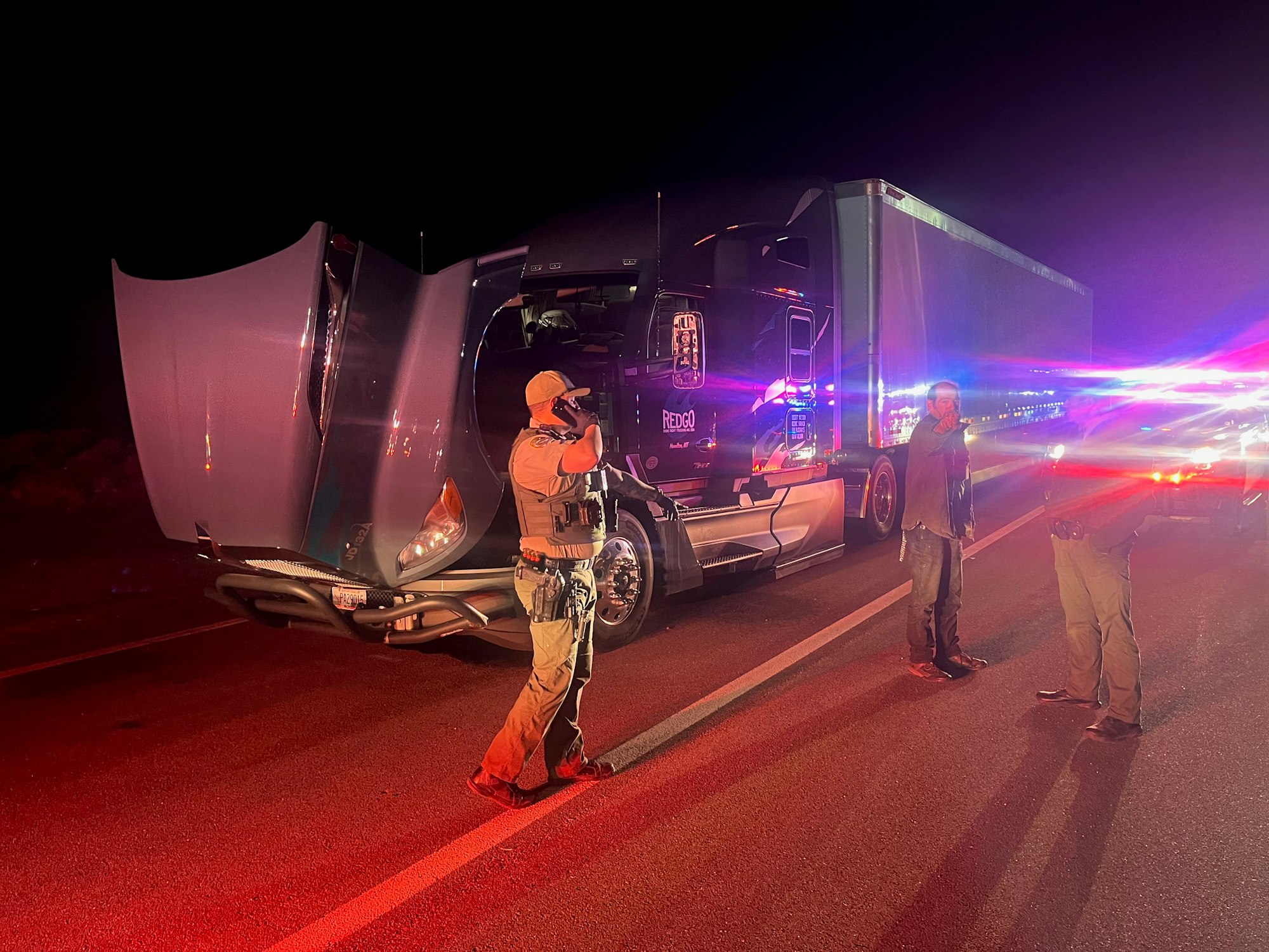 A truck with its hood up, surrounded by law enforcement officers on a road at night, with red and blue lights flashing.