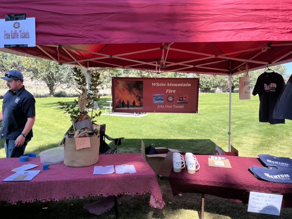 A man stands by a tent with tables displaying merchandise and information, with signs promoting a fire department and recruitment.