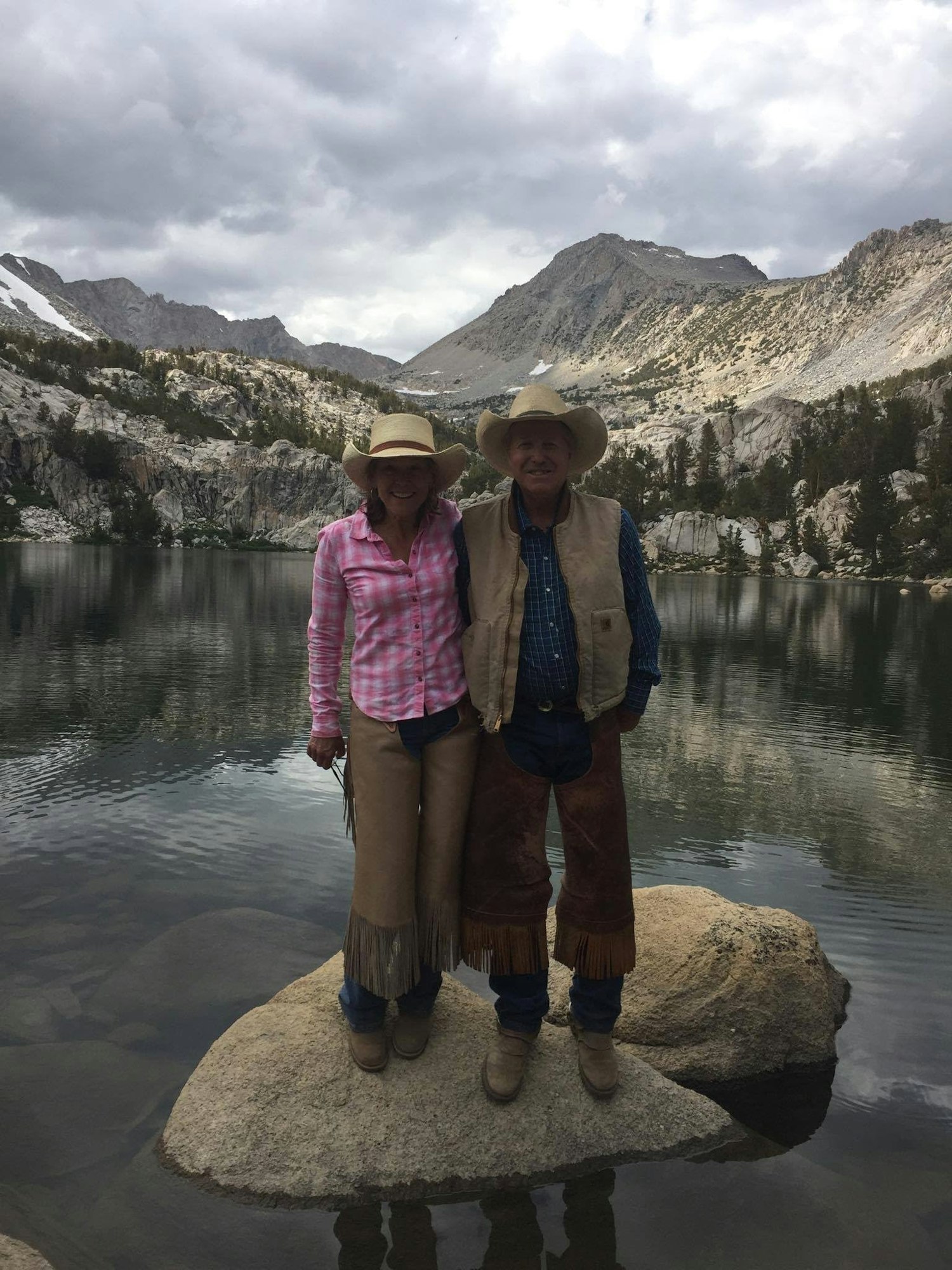 Two people standing on a rock by a mountain lake, wearing cowboy hats and outdoor gear, with overcast skies.