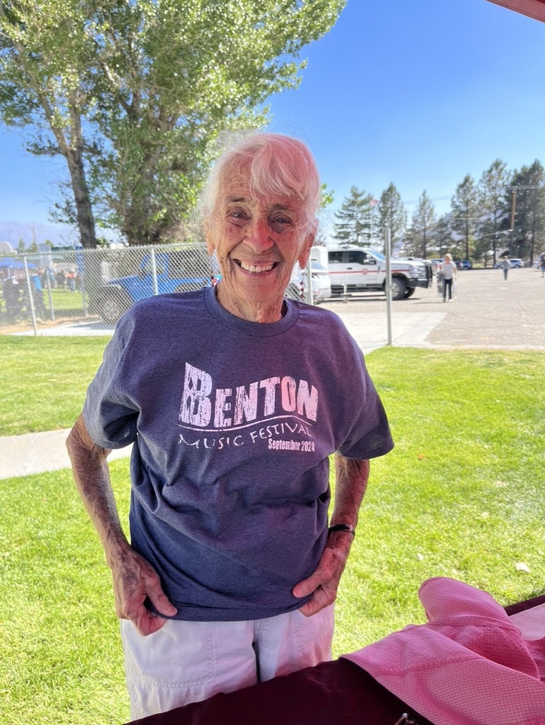Elderly person with a joyful expression, wearing a "Benton Music Festival" T-shirt, outdoors on a sunny day.