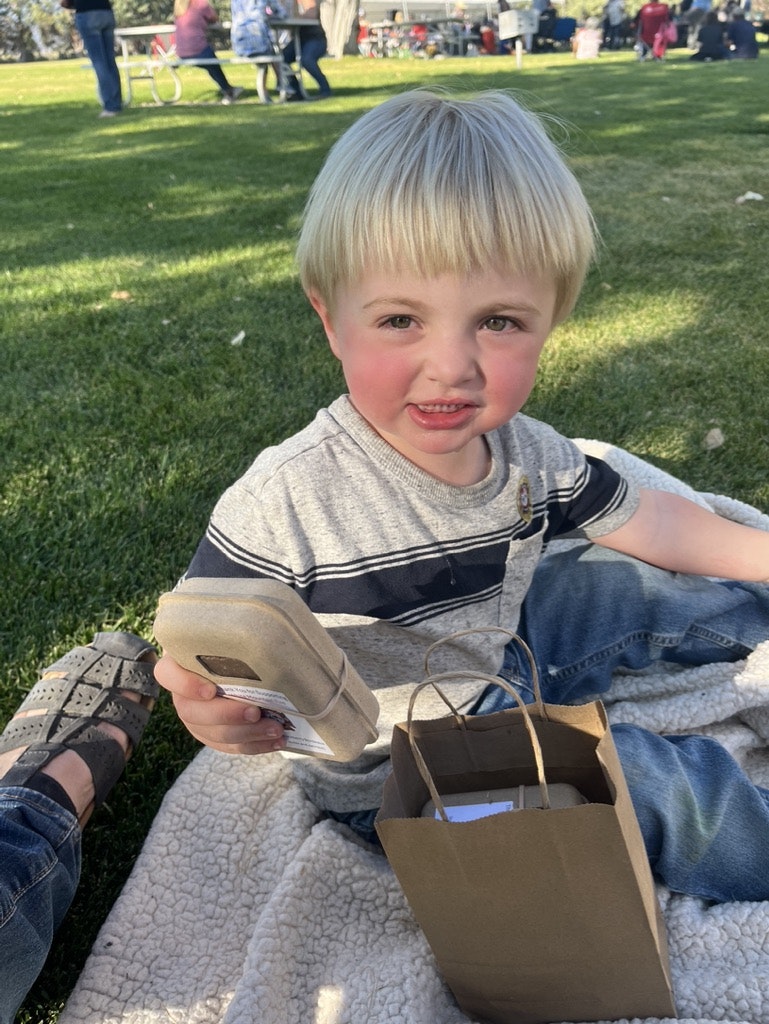 A child sitting on a blanket, a person holding a shoe, a paper bag, outdoors.