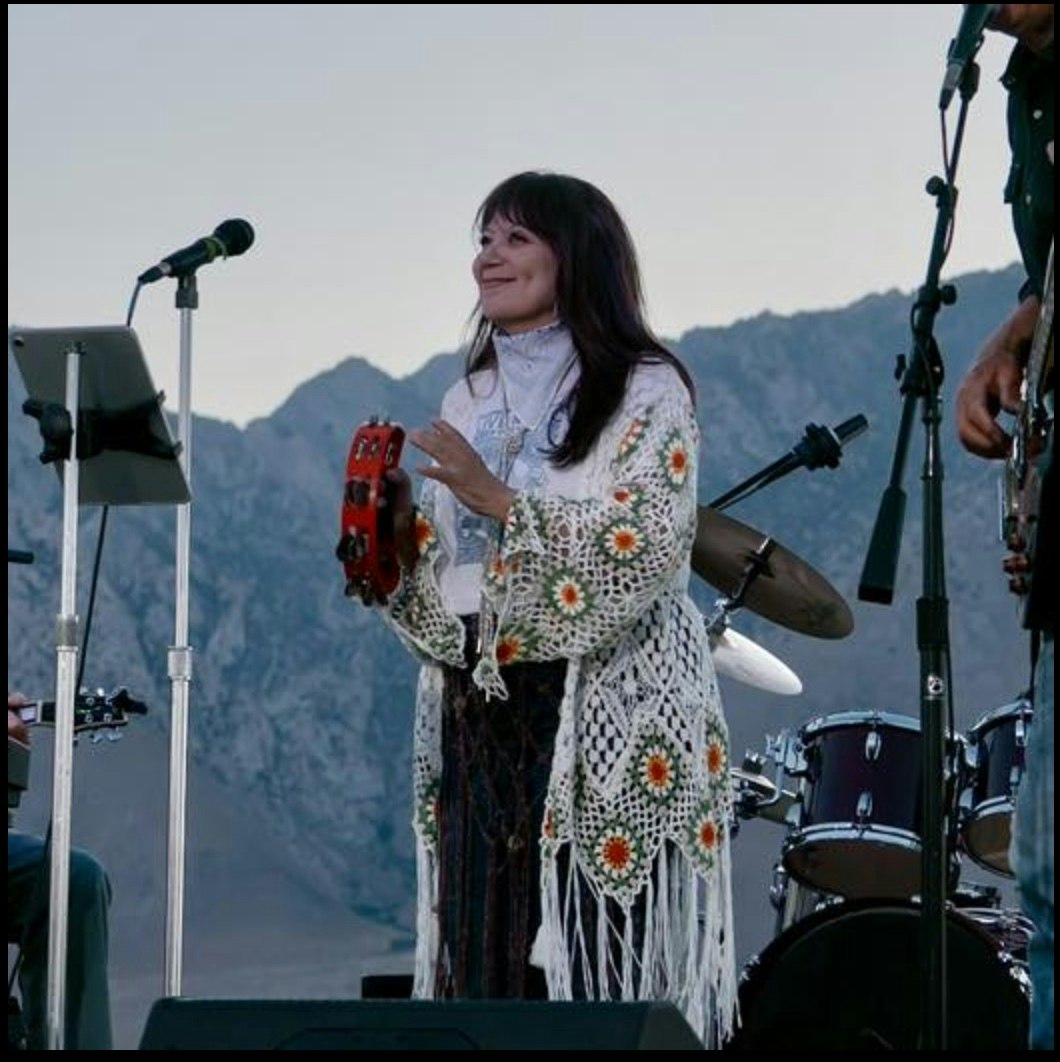 A person is playing a tambourine on stage with a microphone, mountain background, and music equipment visible.
