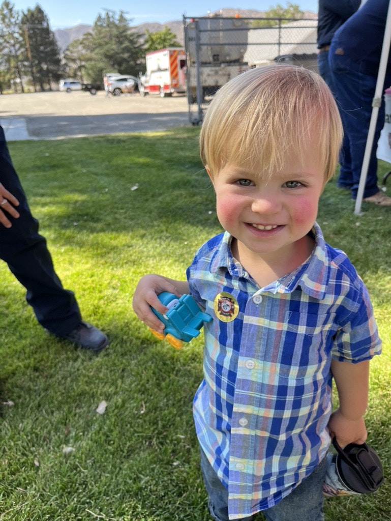 A young child with a toy, wearing a plaid shirt, outdoors with vehicles in the background.