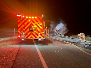 A nighttime scene with firefighters, a firetruck, and an overturned vehicle on a road.