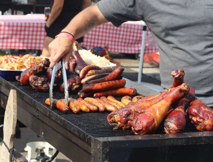 Man grilling assorted sausages and turkey legs, with a dish of vegetables in the background.