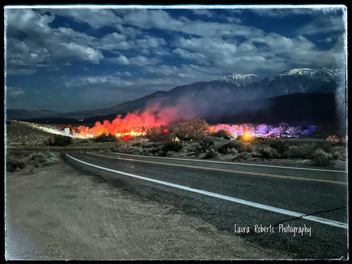 A nighttime scene with a road leading to illuminated, colorful flares or lights against a backdrop of mountains under a cloudy sky.