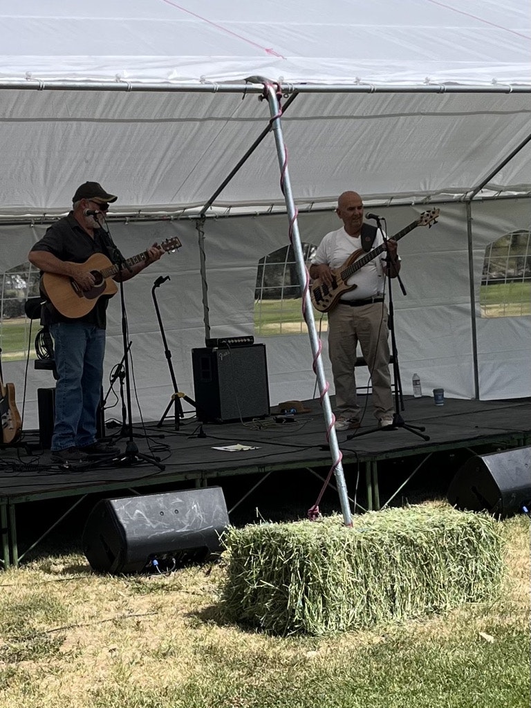 Two musicians playing guitar and bass on an outdoor stage under a tent, with a hay bale in front.