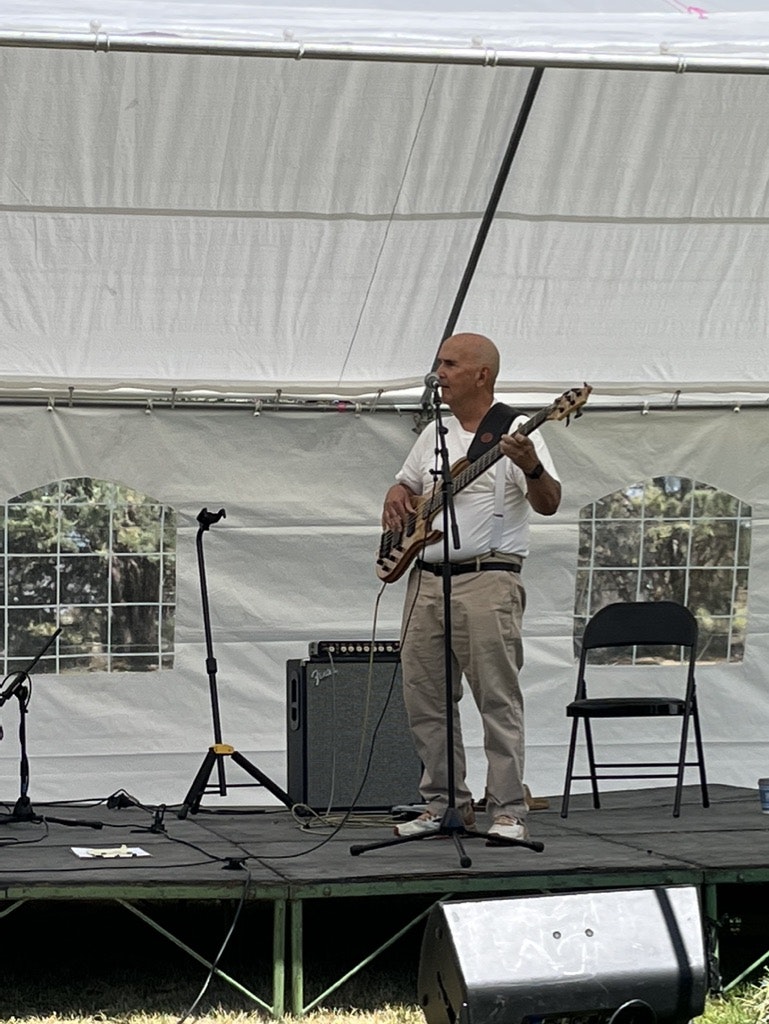A bald man playing a guitar and singing into a microphone on a stage with speakers and a tent-like backdrop.