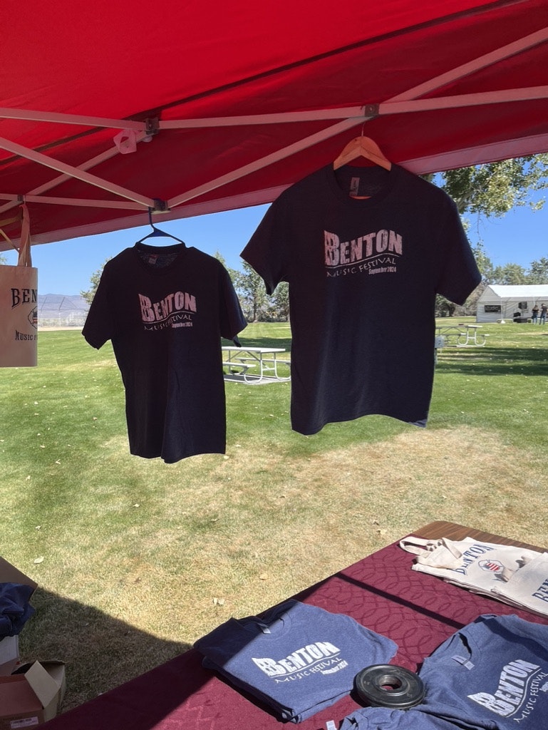 T-shirts hanging under a red canopy at an outdoor event, with "BENTON MUSIC FESTIVAL" printed on them.