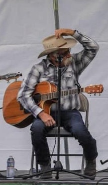A person in a plaid shirt and cowboy hat sits on a chair playing a guitar, with a microphone in front of them.