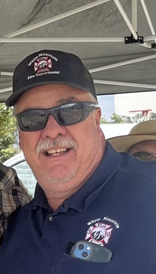 A smiling person wearing sunglasses, a cap, and a shirt with fire department logos, with a cell phone in a pocket.