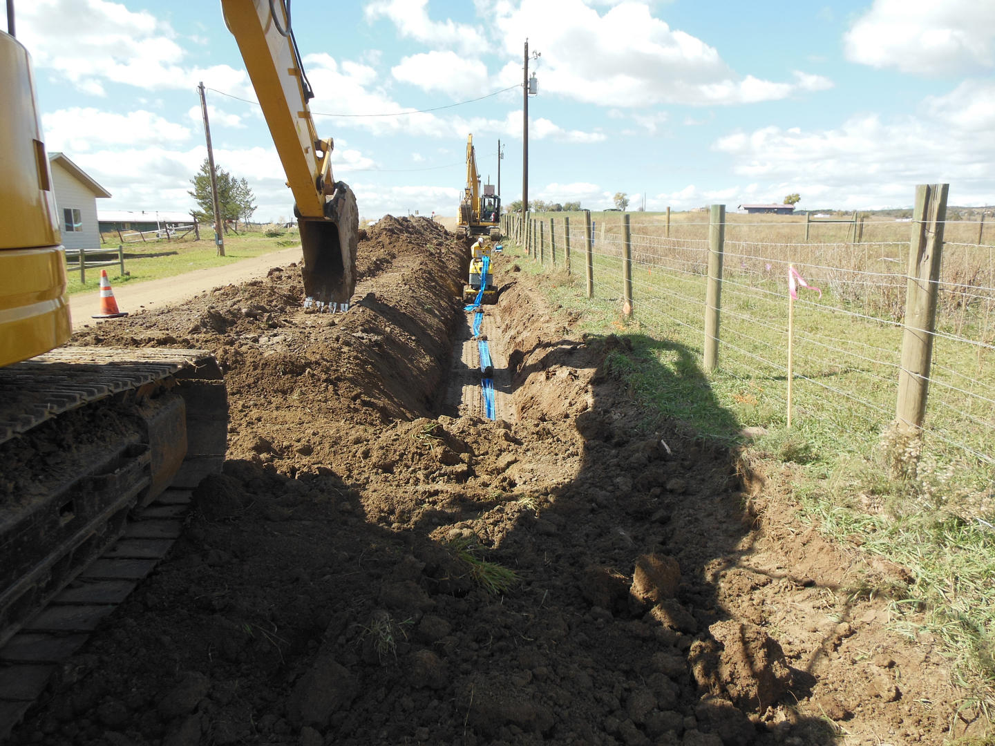 An excavator digging a trench by a fence in a grassy field.