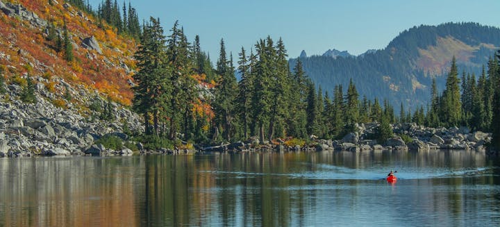 A serene lake surrounded by colorful autumn foliage and evergreen trees, with a person kayaking in a red kayak.