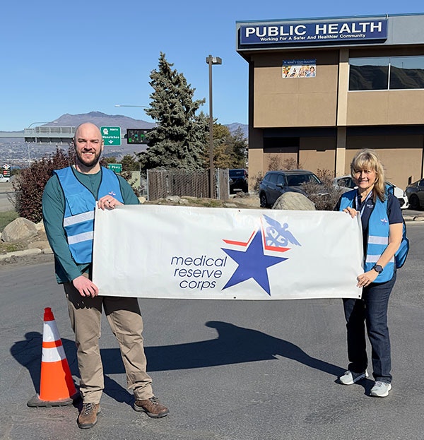 Two volunteers hold a "Medical Reserve Corps" banner outside a public health building, promoting community health initiatives.