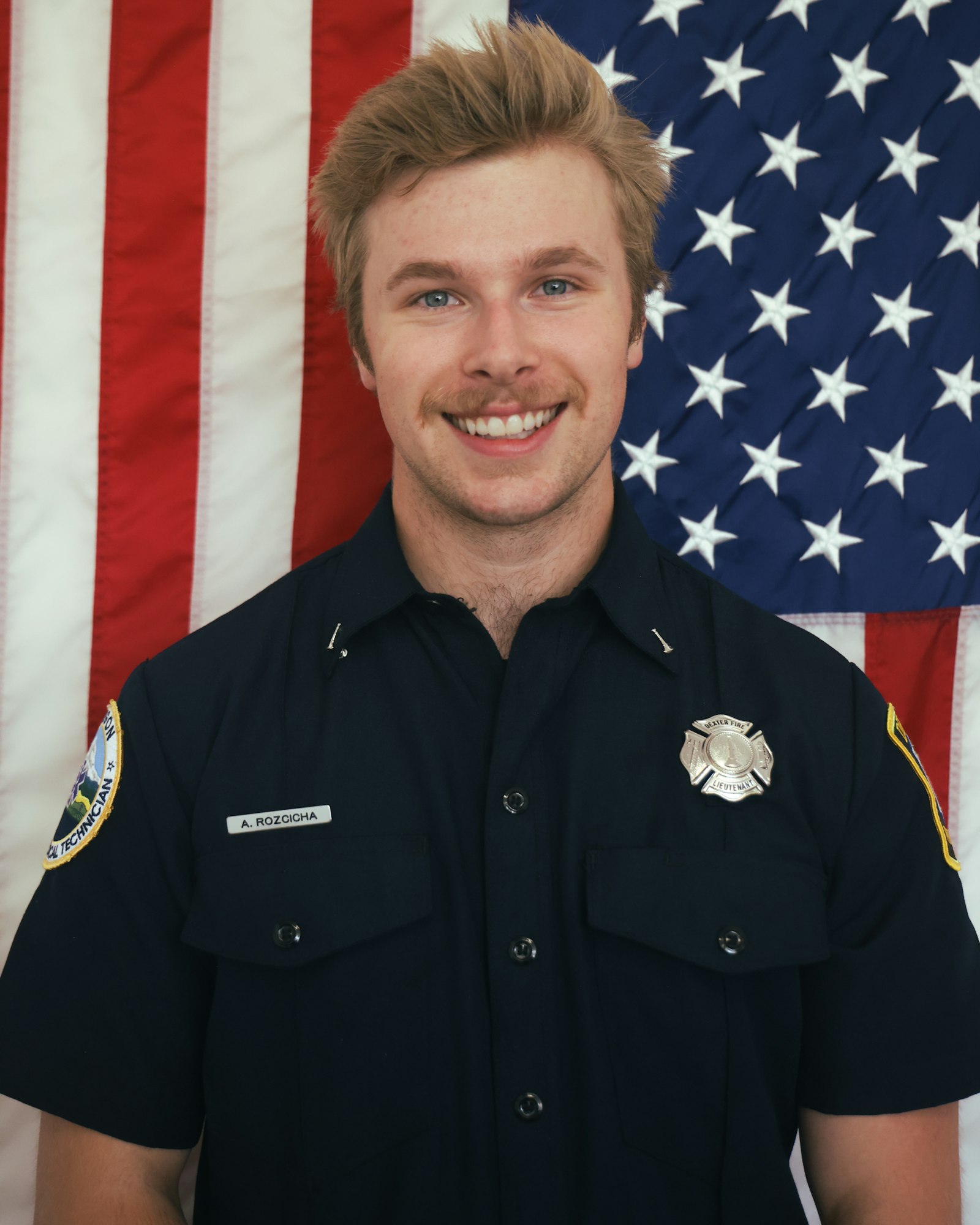 A smiling person in a dark uniform stands in front of a U.S. flag.