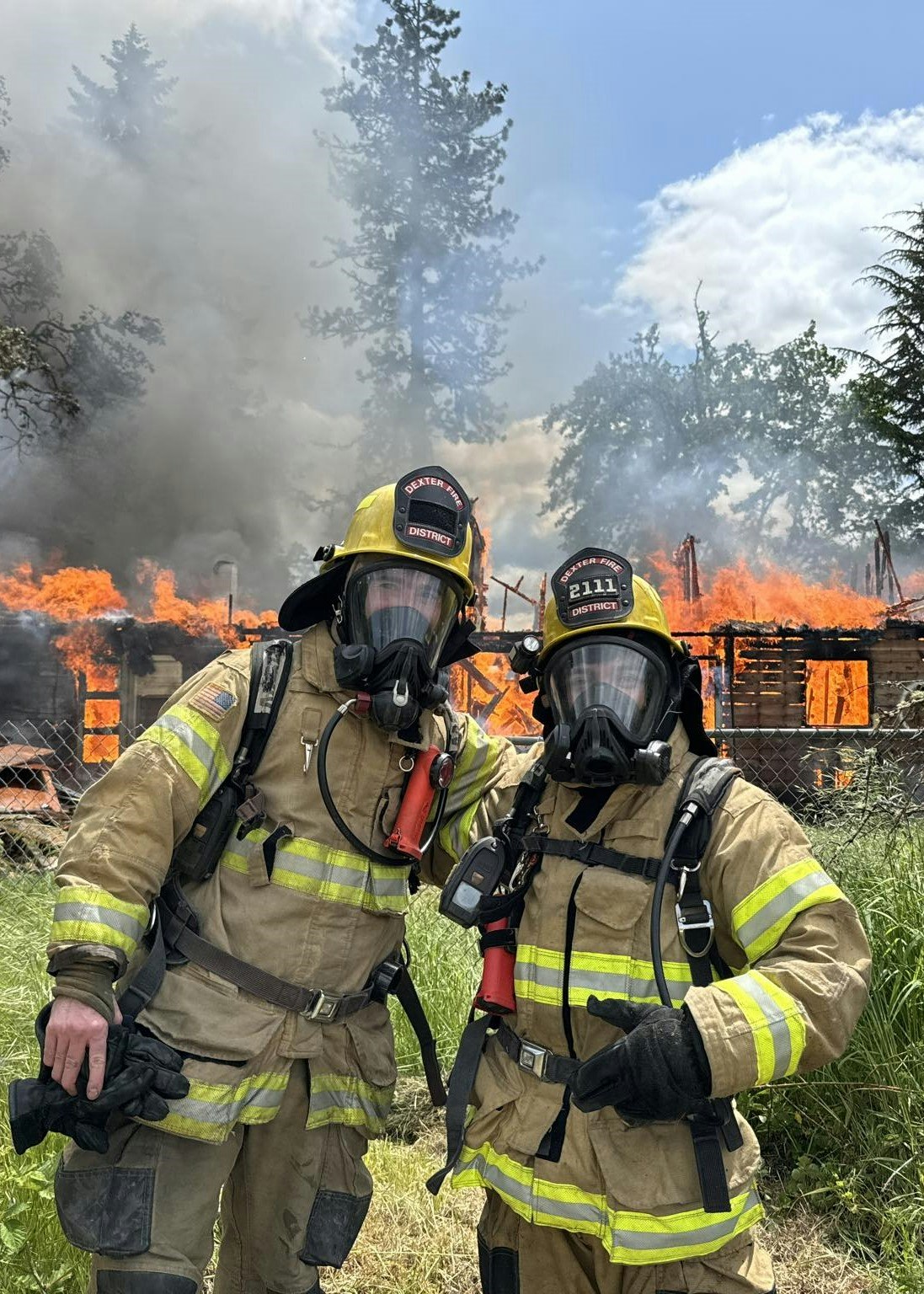 Two firefighters in gear stand in front of a burning building with smoke and flames in the background.