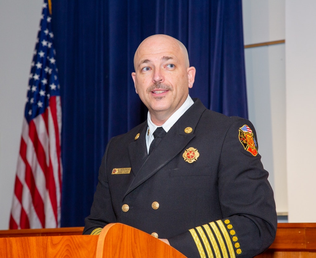 Man in firefighter uniform with badge speaking at podium, American flag in background.