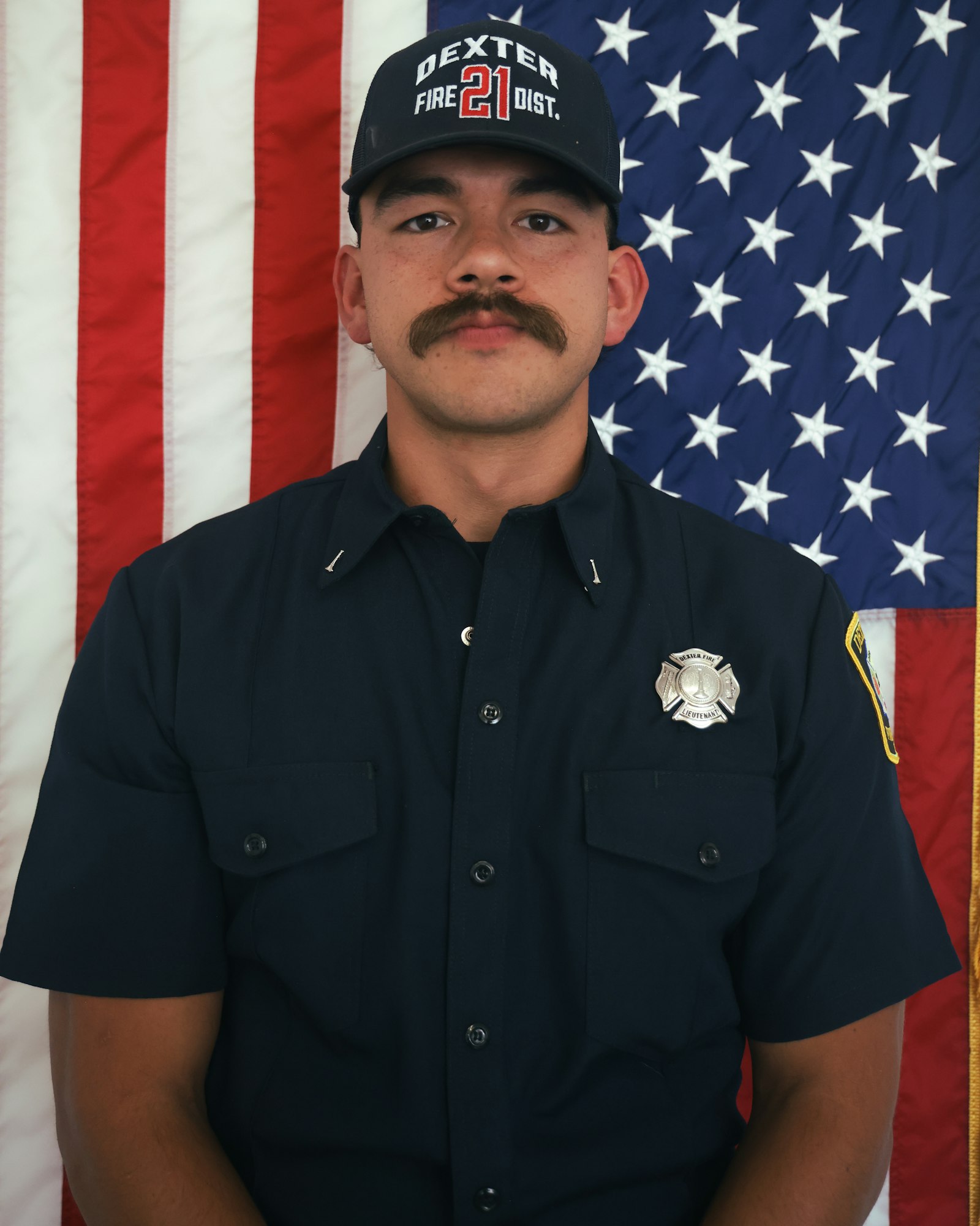A person in a fire department uniform stands in front of an American flag.