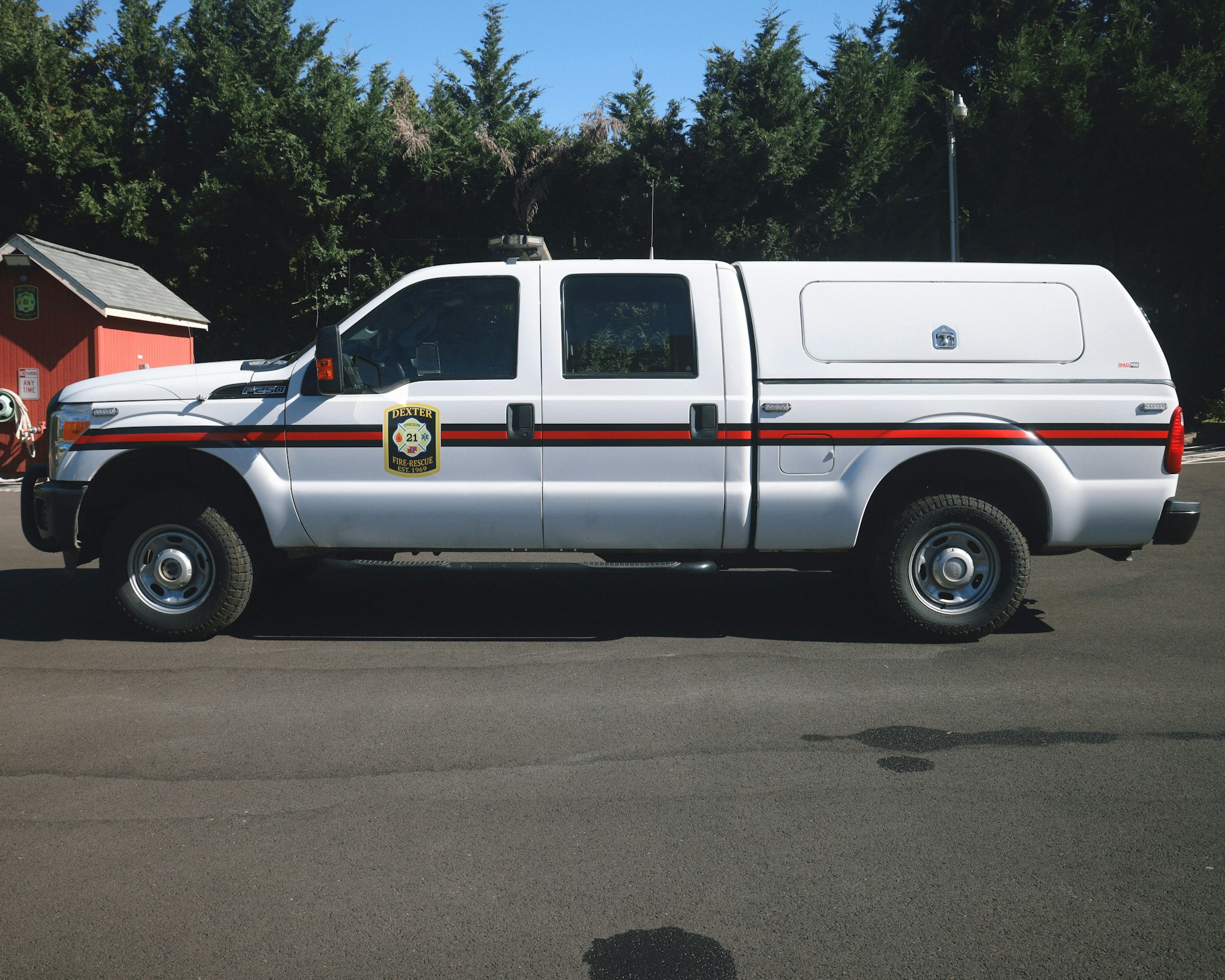 A white fire rescue pickup truck parked on a road in front of a red building, trees in the background.