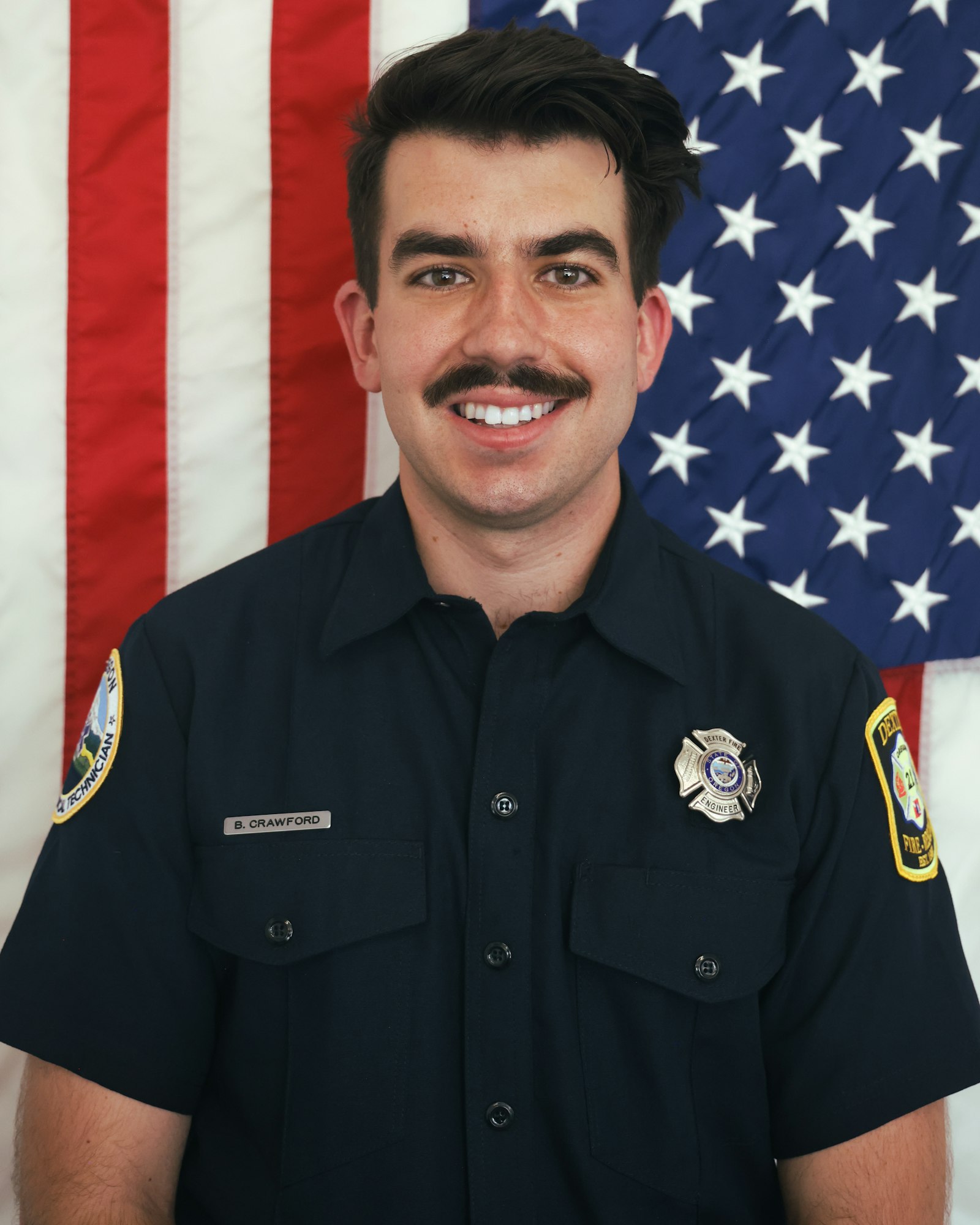 A person in a uniform with badges, standing in front of an American flag.