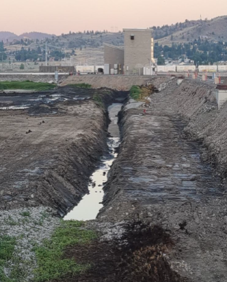 The image shows a barren, muddy landscape with a narrow water channel and a building in the background, surrounded by bare ground and hills.
