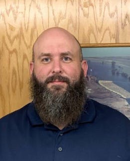 A bearded person in a blue shirt against a wood-paneled wall with a picture frame behind.