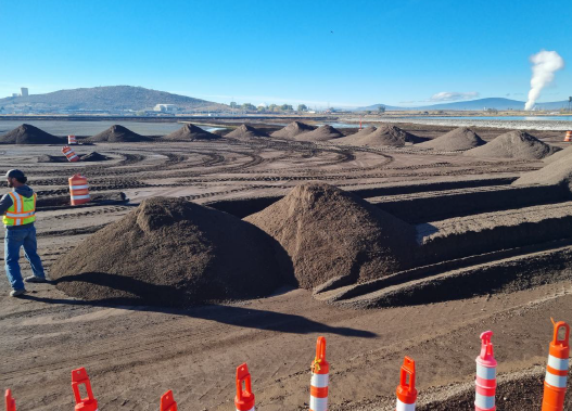 The image shows an area with dirt mounds, a worker in a safety vest, and construction cones, under a clear blue sky.