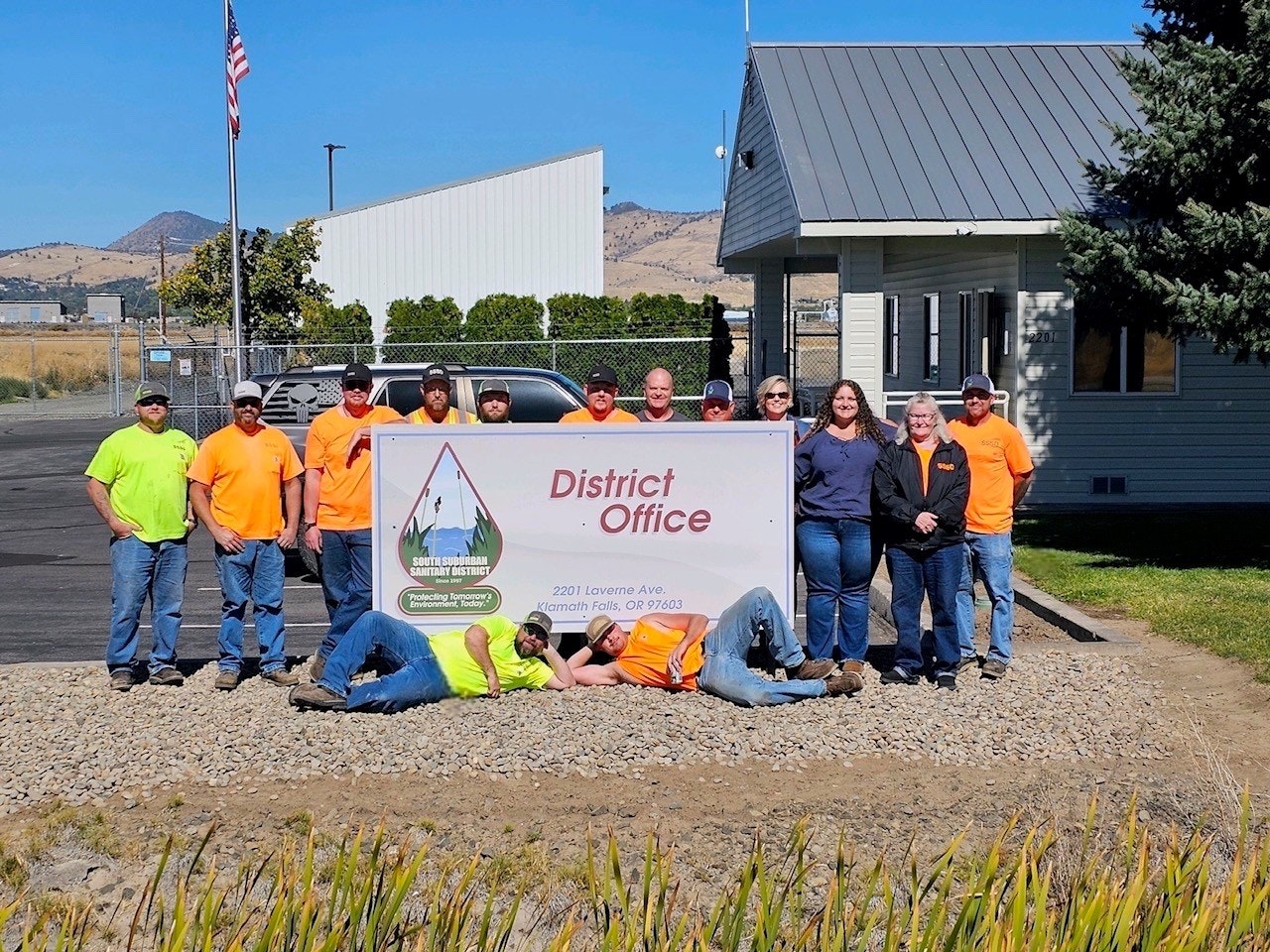 Group of people posing by a "District Office" sign, office building and flag in the background, sunny day.