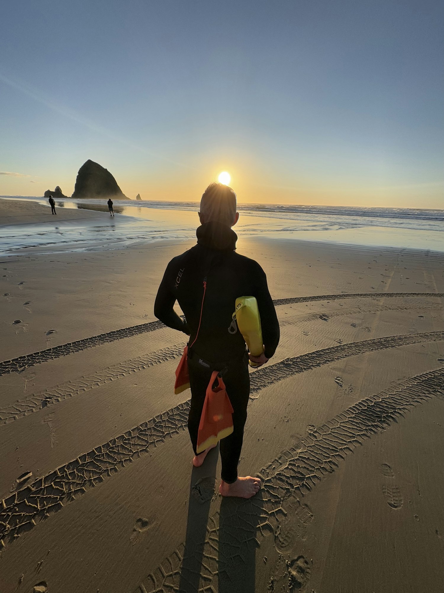 A person in a wetsuit stands on a beach at sunset, holding buoyancy devices, with waves and rocky outcrops in the background.