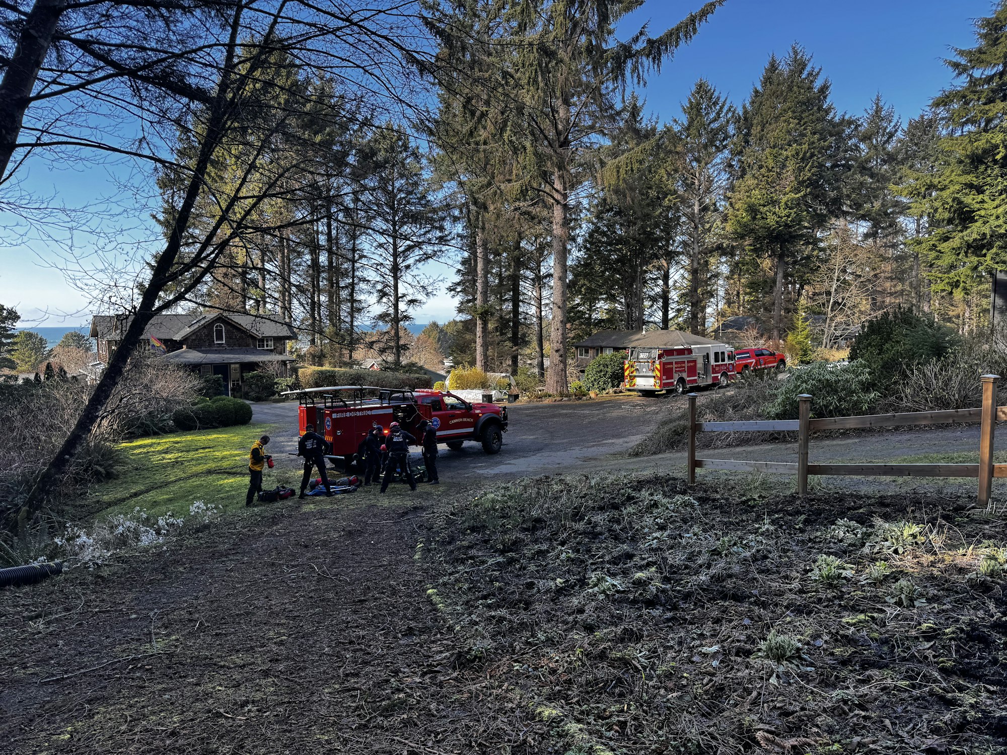 Emergency responders are assisting at a scene near a house, with fire trucks parked nearby in a forested area.