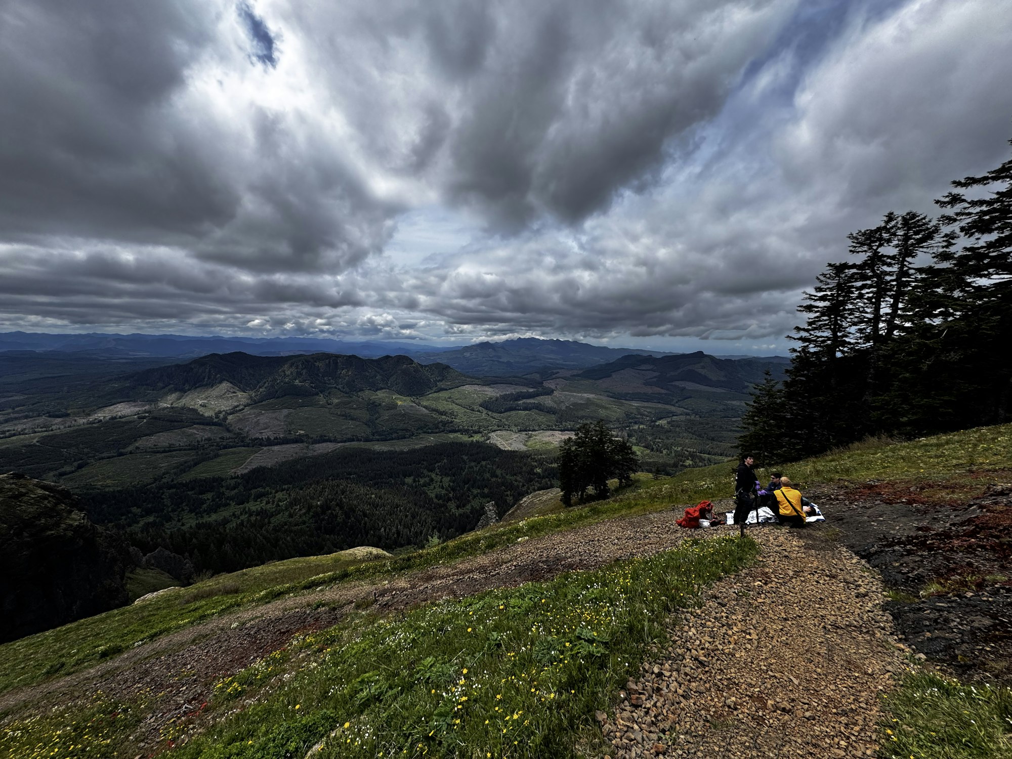 A scenic mountain view under a cloudy sky, with two people seated on the ground, surrounded by lush greenery and distant hills.