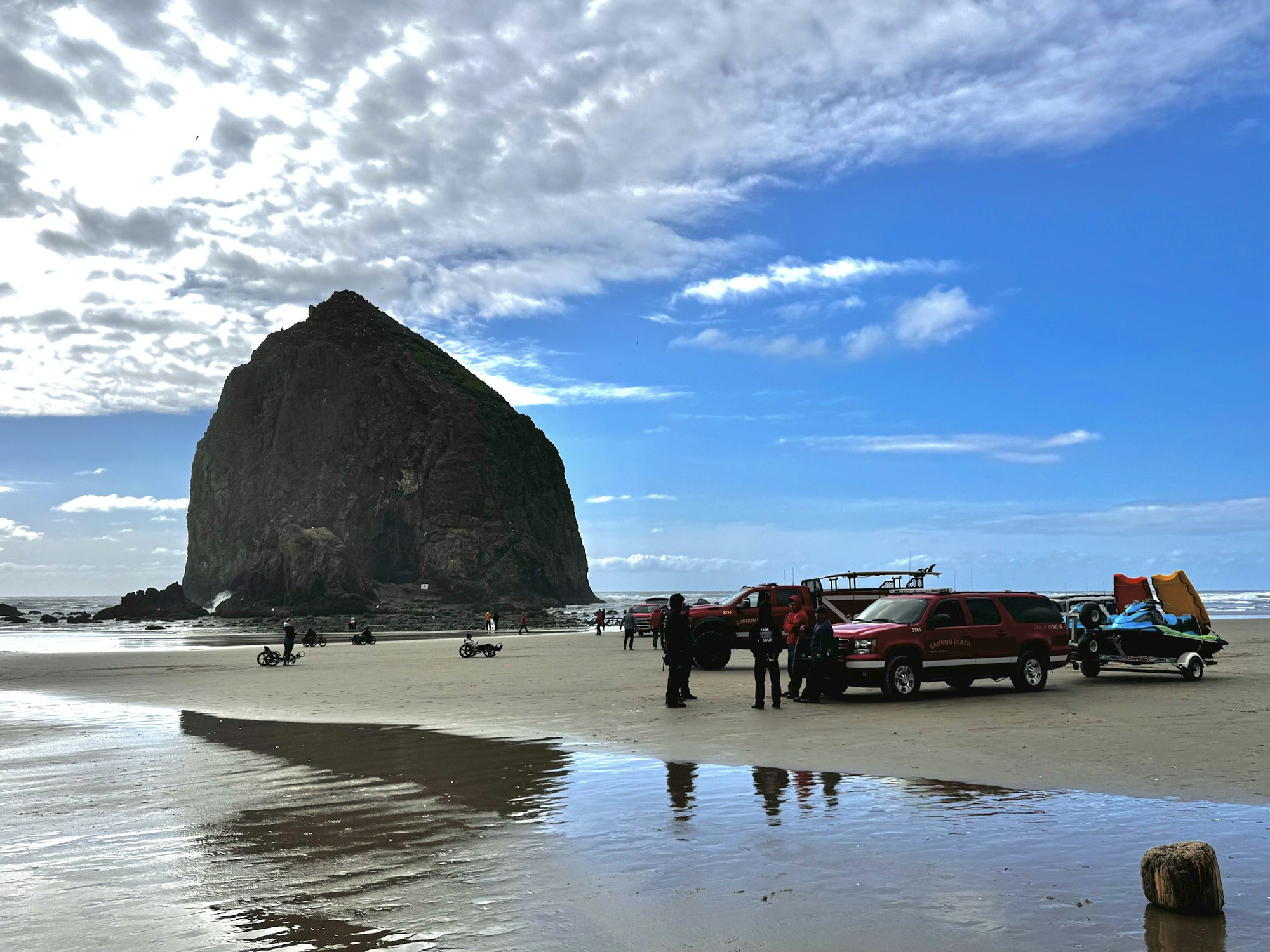 A beach scene features a large rock formation, vehicles, and people engaging in activities with watercraft under a cloudy sky.