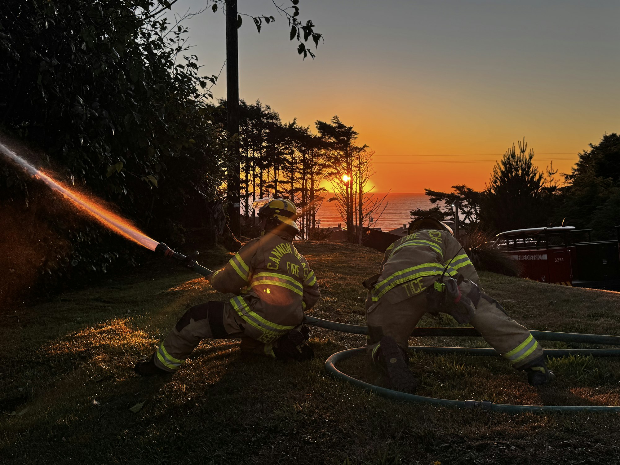 Two firefighters spray water as the sun sets over the ocean, creating a dramatic scene with vibrant colors.