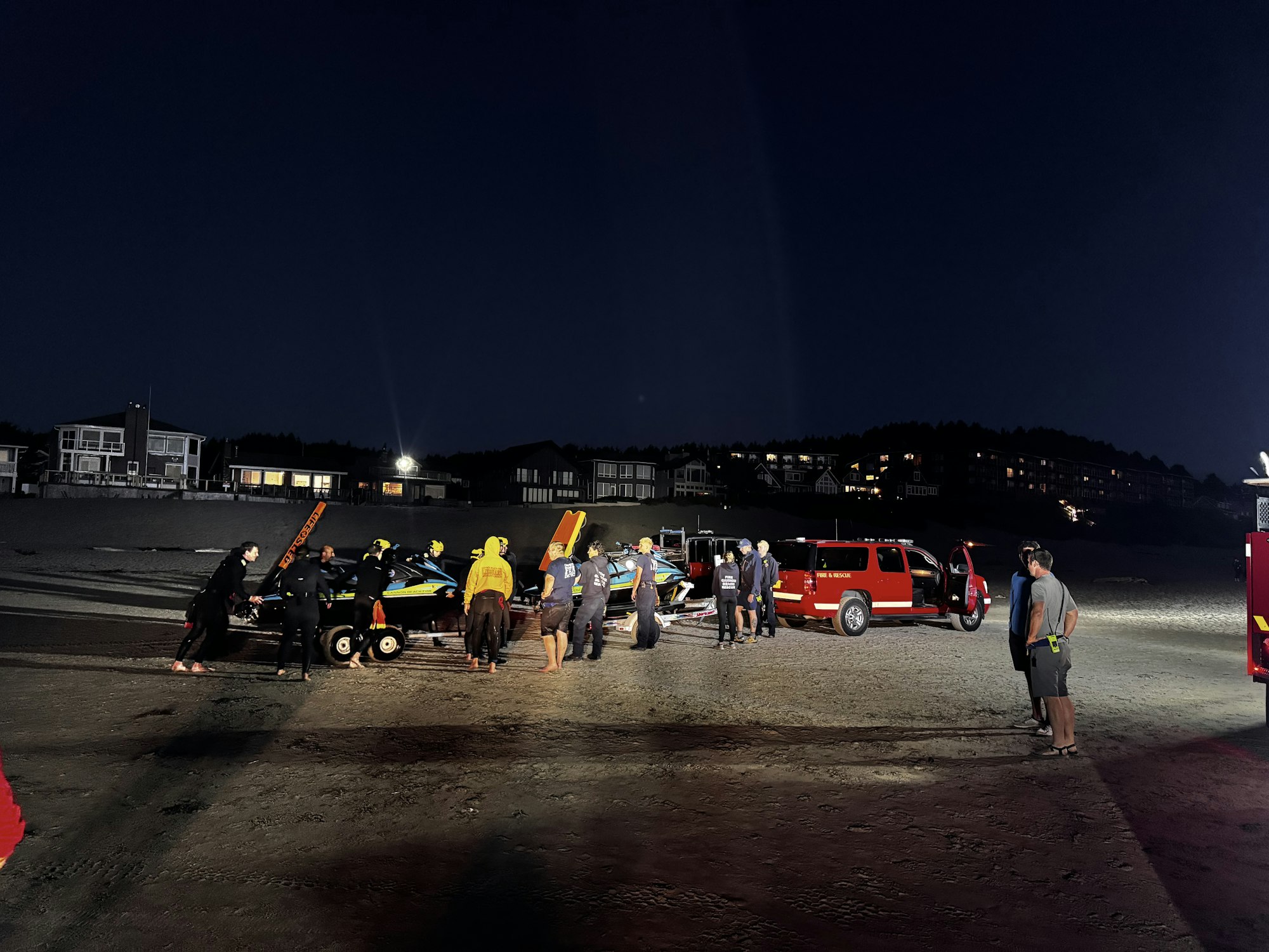 A group of people, possibly rescue teams, are gathered on a beach at night, with vehicles and equipment present.