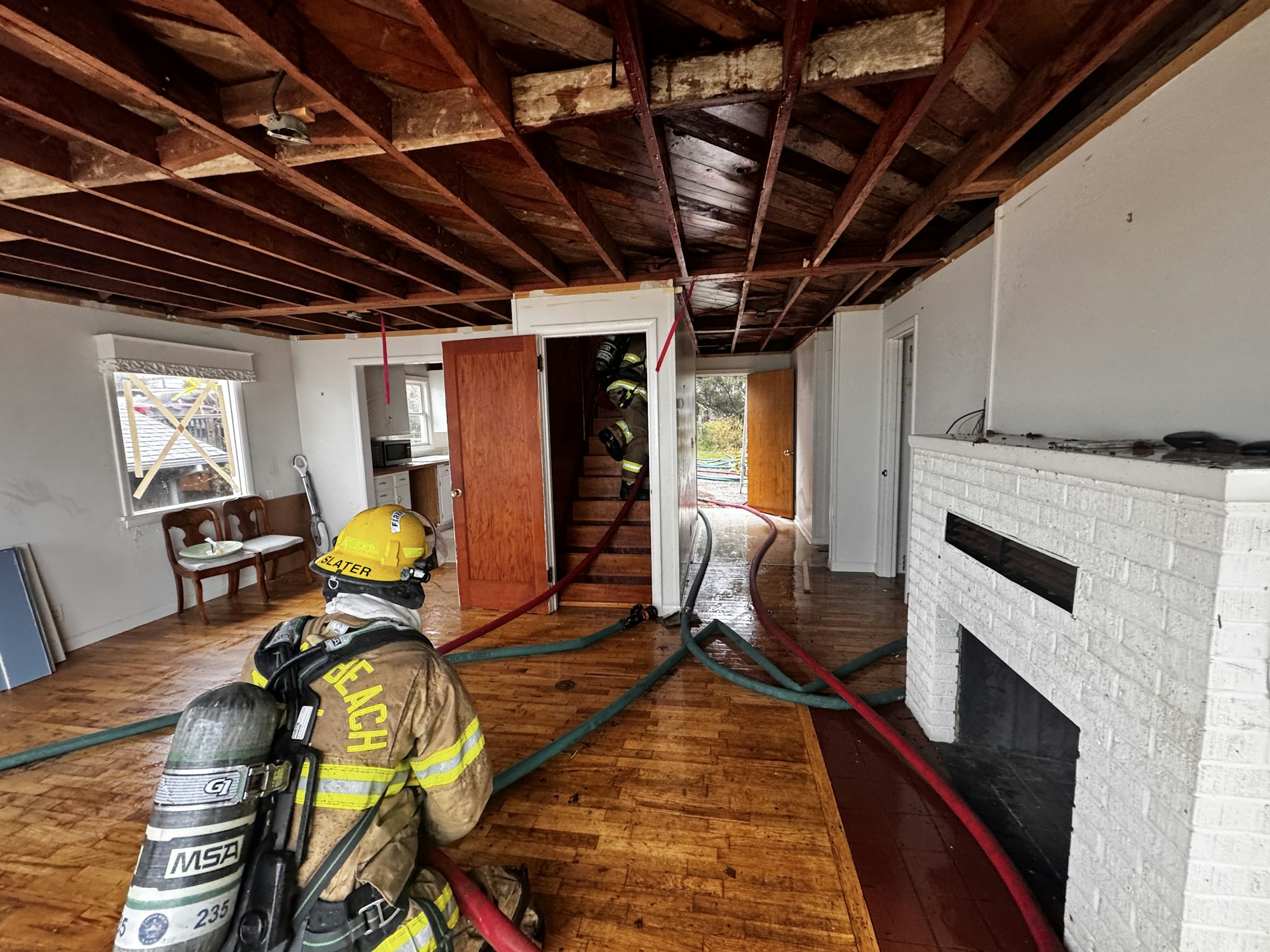 A firefighter prepares in a water-damaged home, with hoses laid across the floor and another firefighter visible in the background.