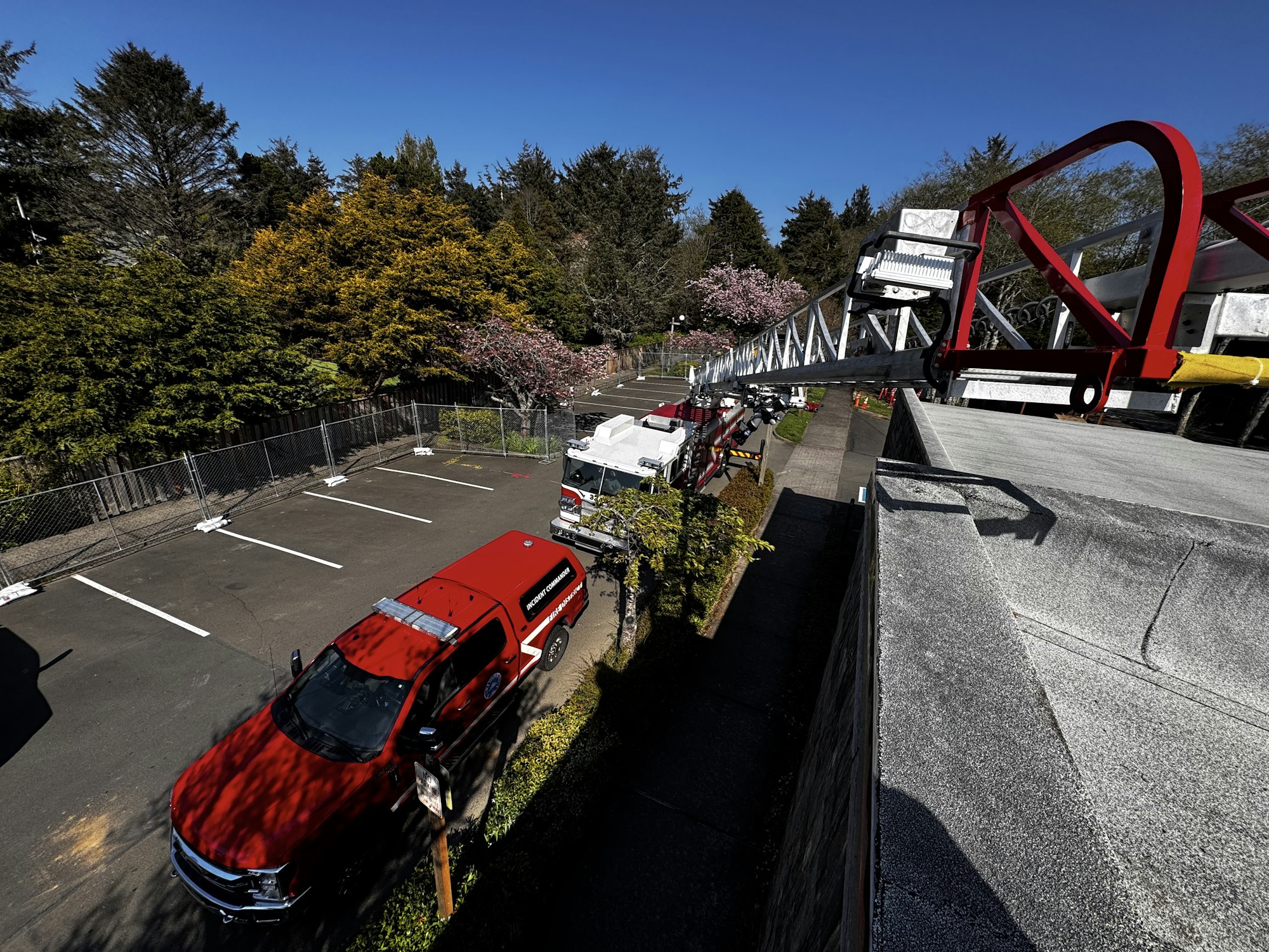 The image shows a red fire truck and ladder, parked near trees and a fenced area, under a clear blue sky.
