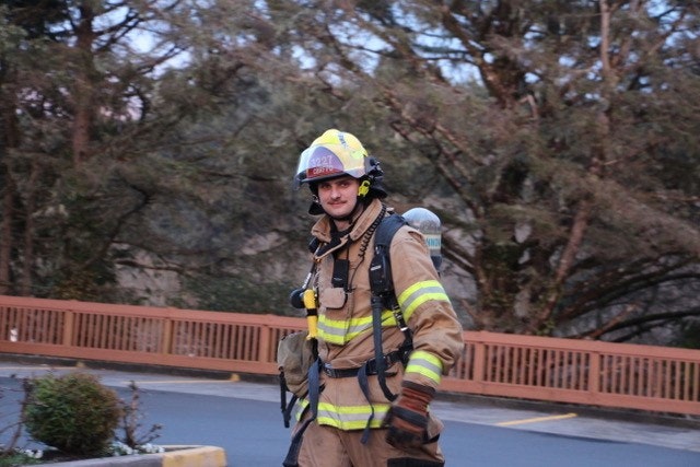 A firefighter in gear with a helmet walks outdoors, surrounded by trees, displaying readiness and confidence.