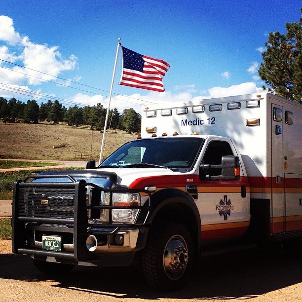 An ambulance labeled "Medic 12" parked under an American flag, surrounded by a natural landscape with trees and blue skies.